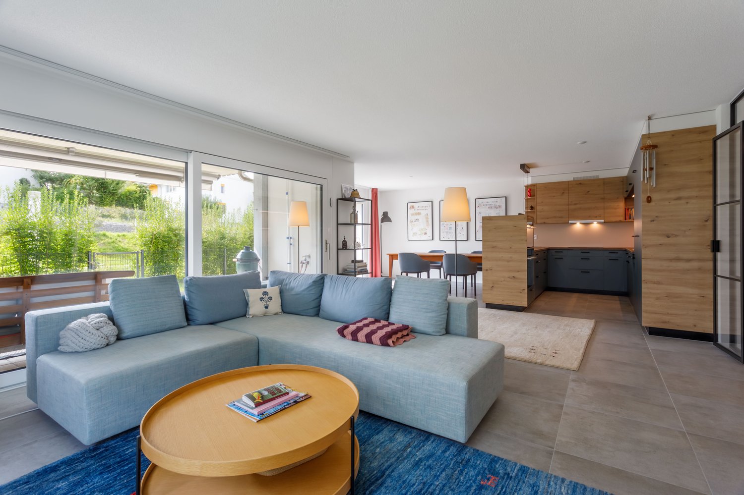 Blue couch, large glass windows, wooden table, rug, kitchen with dining table and chairs, wooden cabinets.