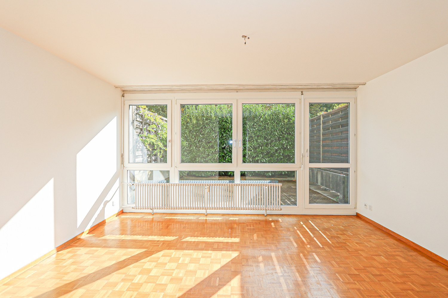 Spacious, empty room with large windows and balcony access, wooden floor, white walls, built-in radiator, and a garden view.