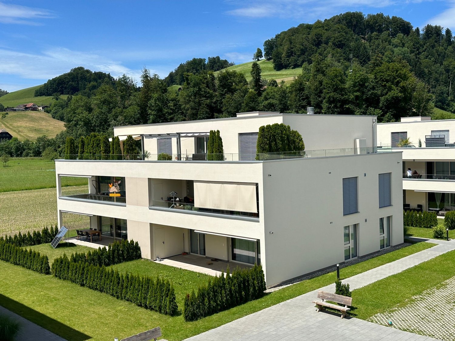 A modern, multi-story apartment building with a white exterior and large windows. The building has balconies on some of the units and is surrounded by a well-manicured lawn and landscaping. The building is situated in a scenic, hilly area with trees and g