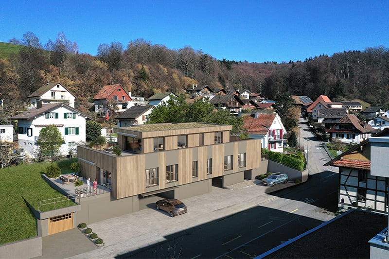 A modern multi-story building with a parking lot in the foreground, surrounded by a residential neighborhood with traditional houses and buildings in a hilly, forested area.