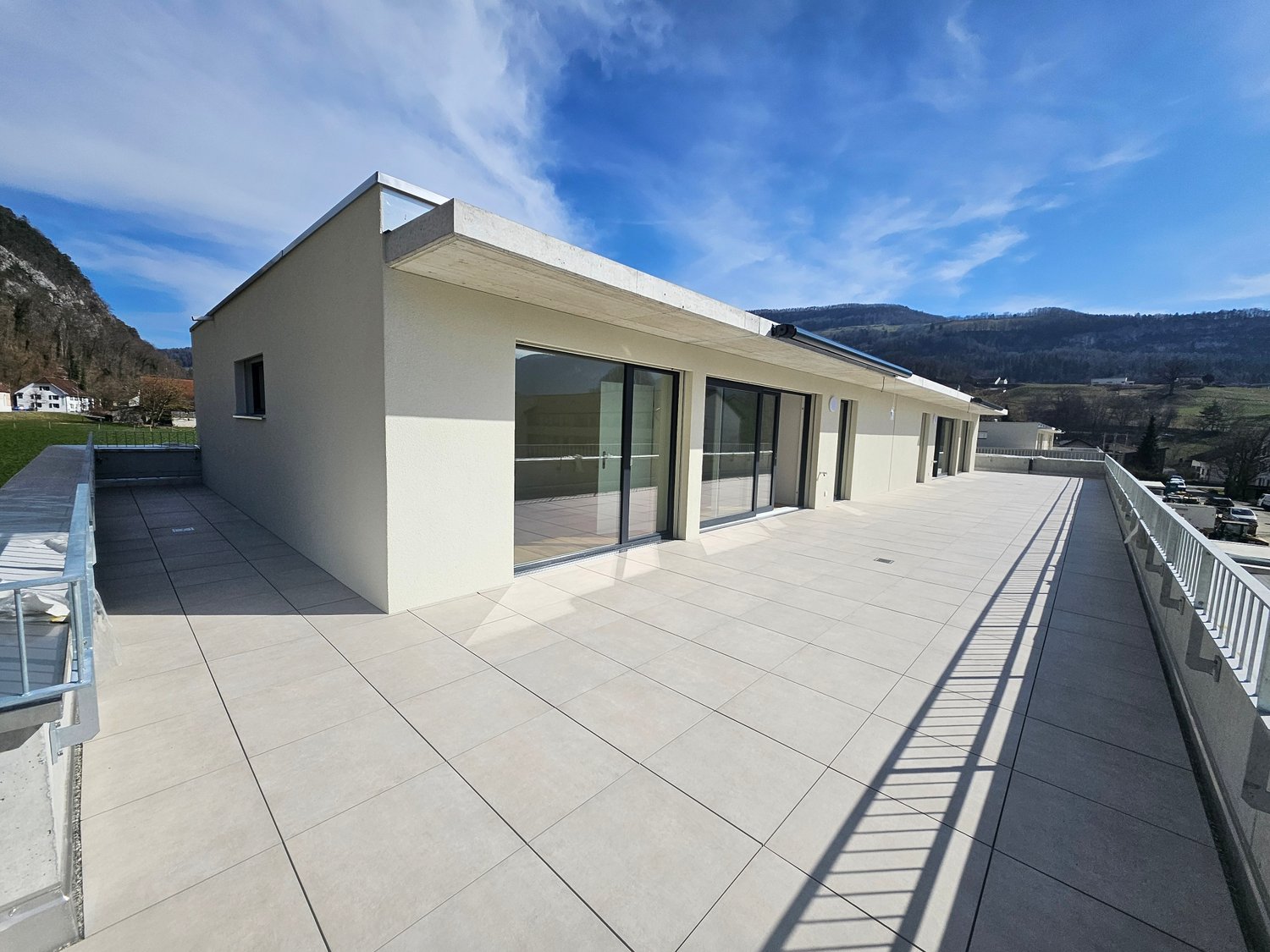 A modern, two-story building with a flat roof and large glass windows. The building has a spacious balcony or terrace area with a railing. The exterior walls are a light, neutral color. The surrounding landscape includes mountains and a clear blue sky.