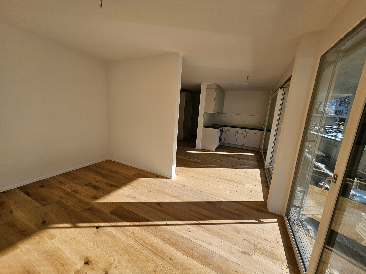 The image shows an empty apartment with hardwood floors, white walls, and an open floor plan. The kitchen area is visible in the background, with white cabinets and appliances. The room has large windows that allow natural light to enter the space.