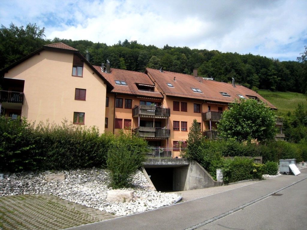 Three-story building, beige exterior, balconies, windows, trees, shrubs, paved road