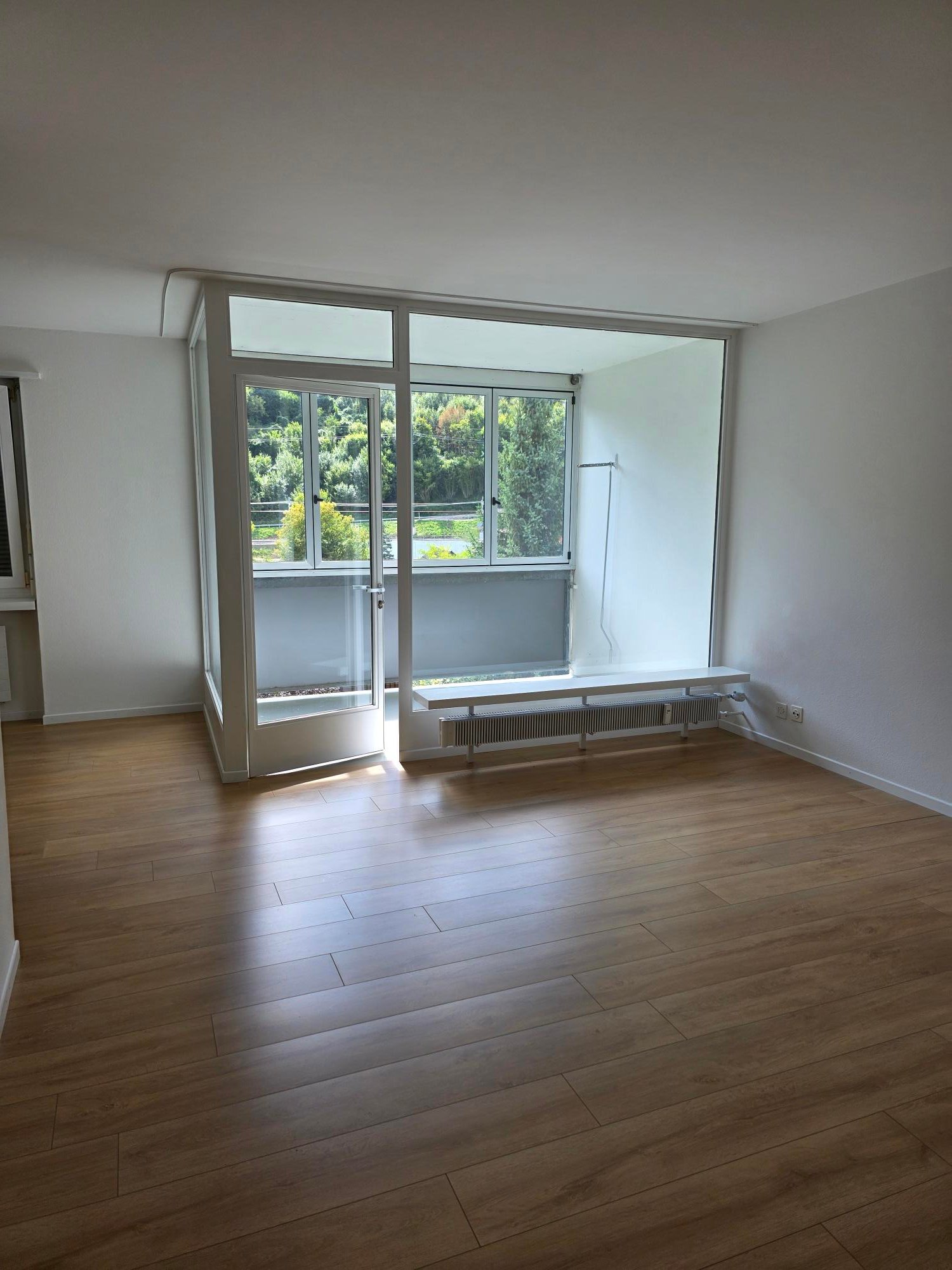 An empty room with sliding glass doors leading to a balcony, wooden flooring, white walls, white benches, and power outlets on the wall.