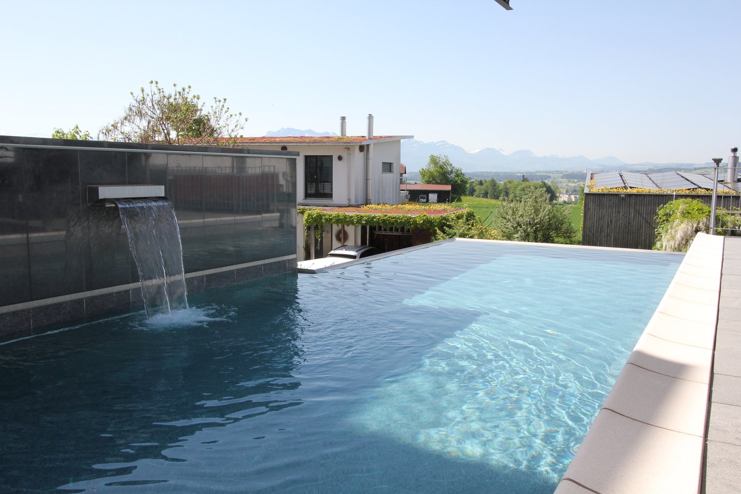 Modern house with a large infinity pool, waterfall feature, and a balcony or terrace overlooking a scenic landscape with mountains in the background.