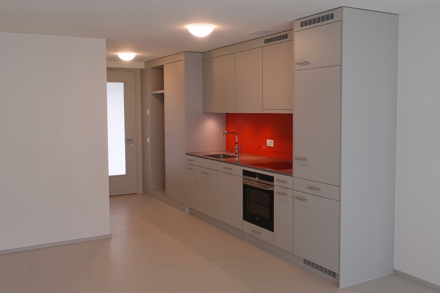 Modern kitchen with white cabinets, red accent wall, built-in oven, stainless steel sink, dishwasher, and white tiled flooring.