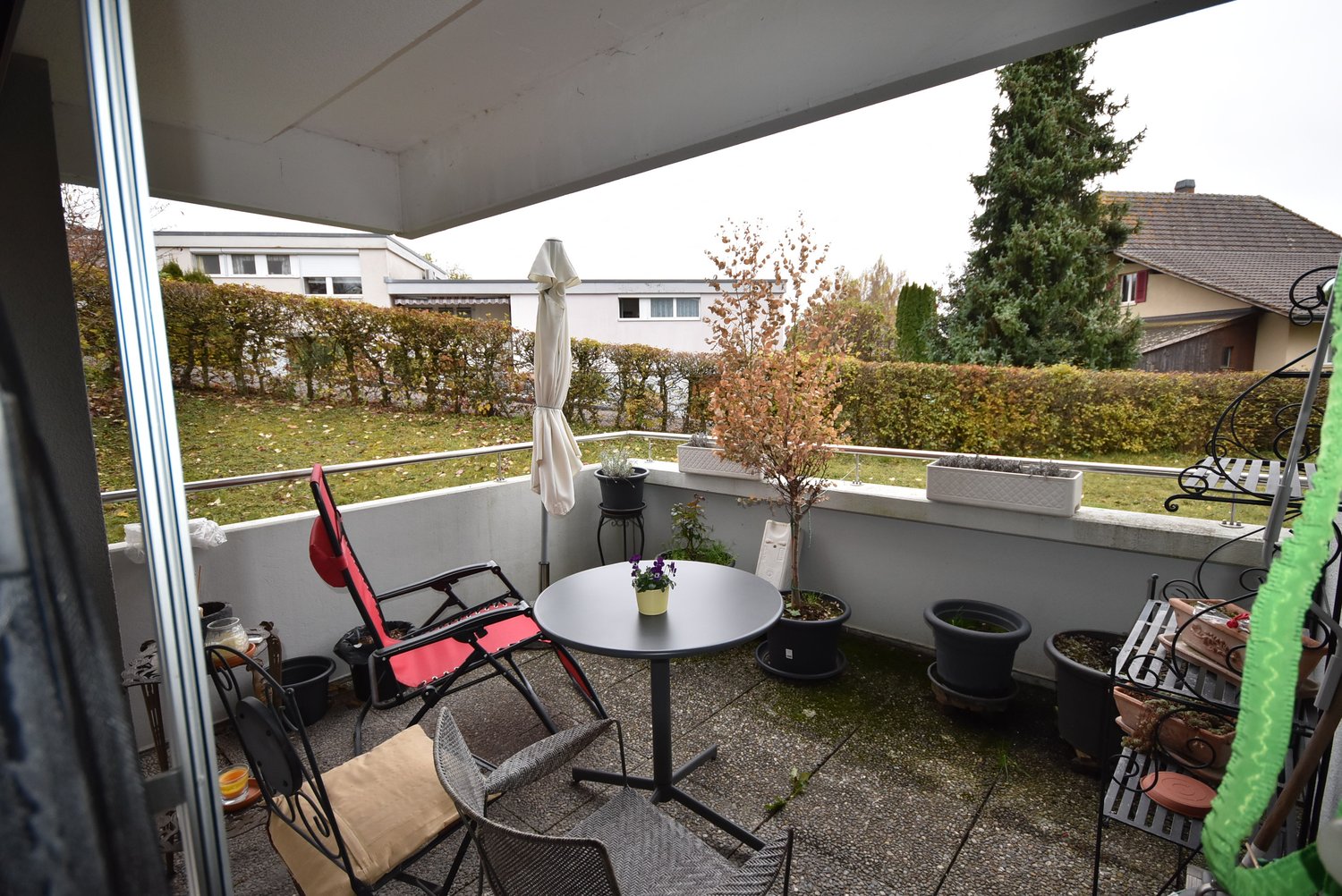 a balcony with two chairs and a table, surrounded by potted plants, a closed umbrella and a metal shelving unit