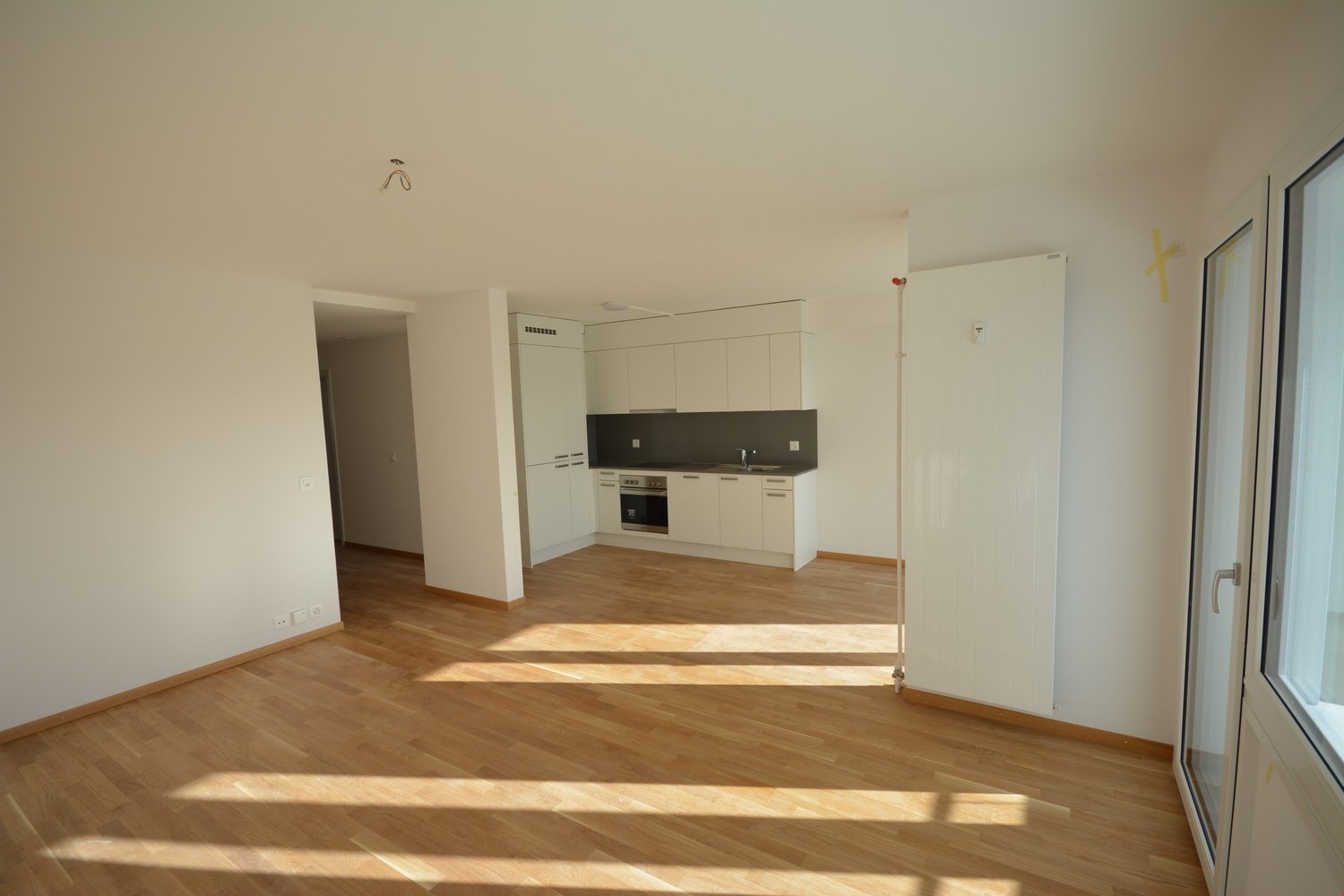 An empty room with wooden floors, white walls, a glass door, kitchen area with oven and sink, cabinets and counters.