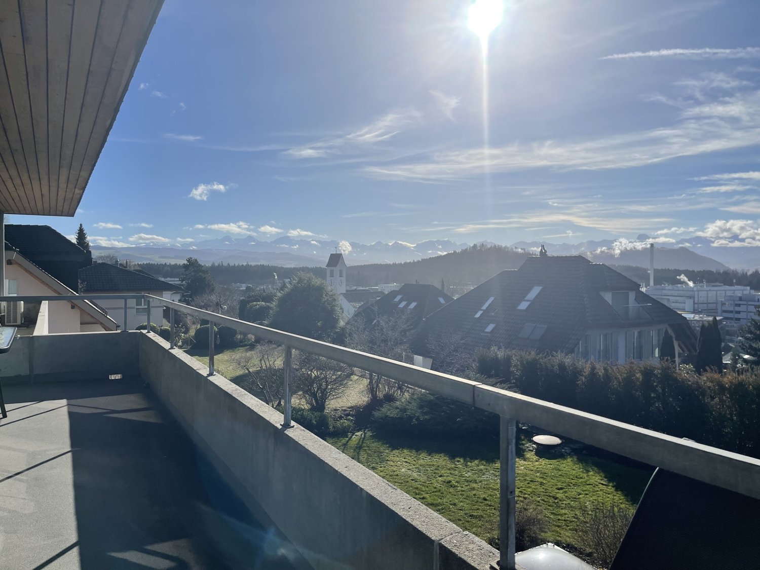 Balcony with metal railings, view of the mountains, sunlight, residential buildings in the background.