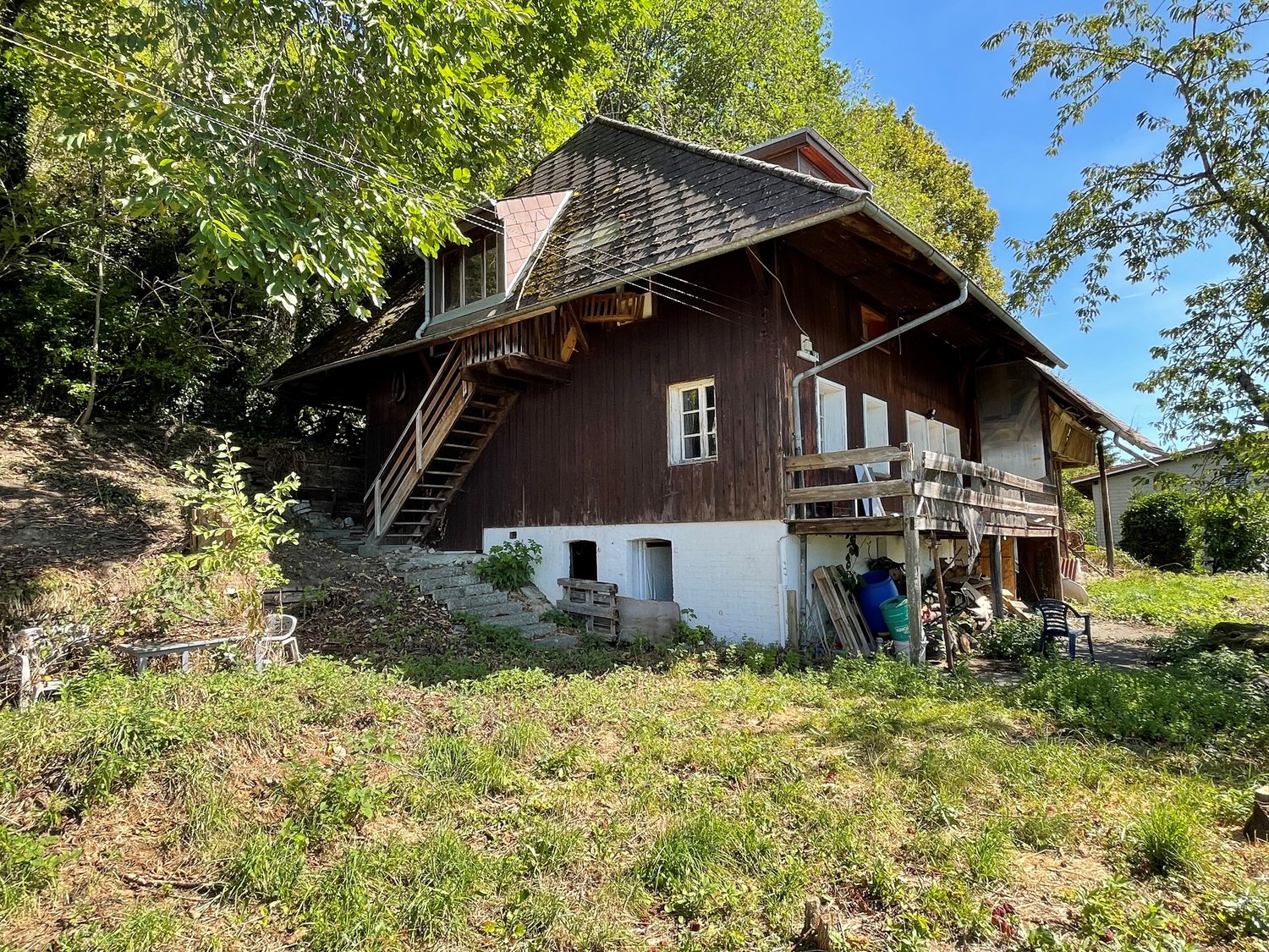 House on a hillside with wooden stairs, wooden porch, brown roof, white windows, white lower part