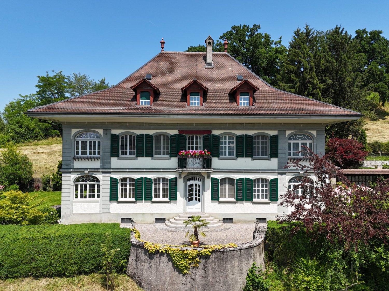 Two-story villa, brown roof, white walls, green shutters, balcony with flower box, entrance with stairs, garden with hedge and fountain, surrounding greenery and trees