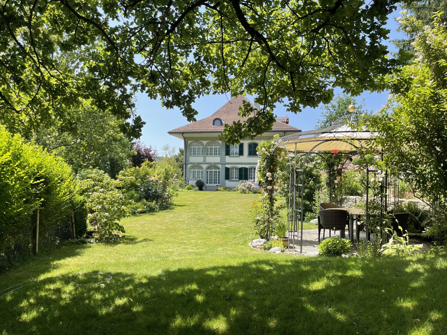 Large house with brown roof, white walls, multiple windows, surrounded by a well-maintained garden with a gazebo, table, and chairs.