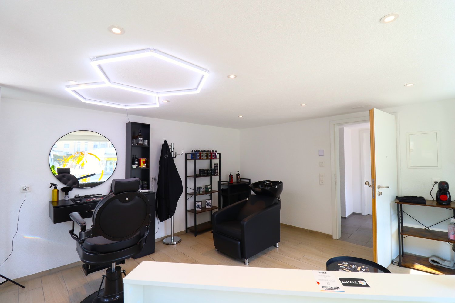 Spacious hair salon with modern decor, including black salon chairs, shelves with hair products, and a large circular mirror. The room has a bright, open layout with recessed lighting fixtures on the ceiling.