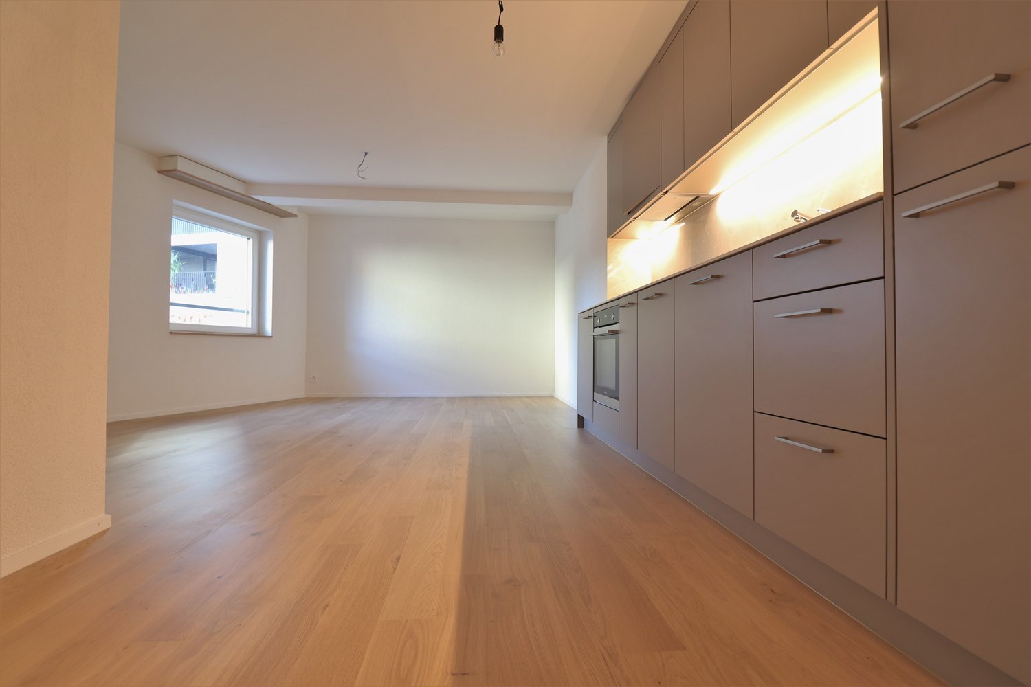 Empty room with wooden floors, white walls, built-in kitchen cabinets, and a window with outside view.