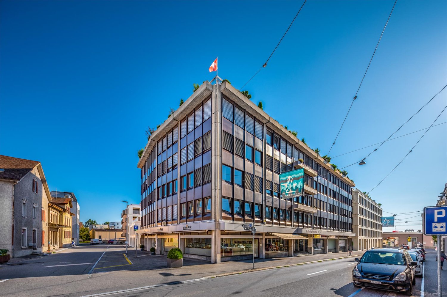 Concrete and glass building, three stories, several windows, signage, parked cars, street, clear sky, electrical cables.