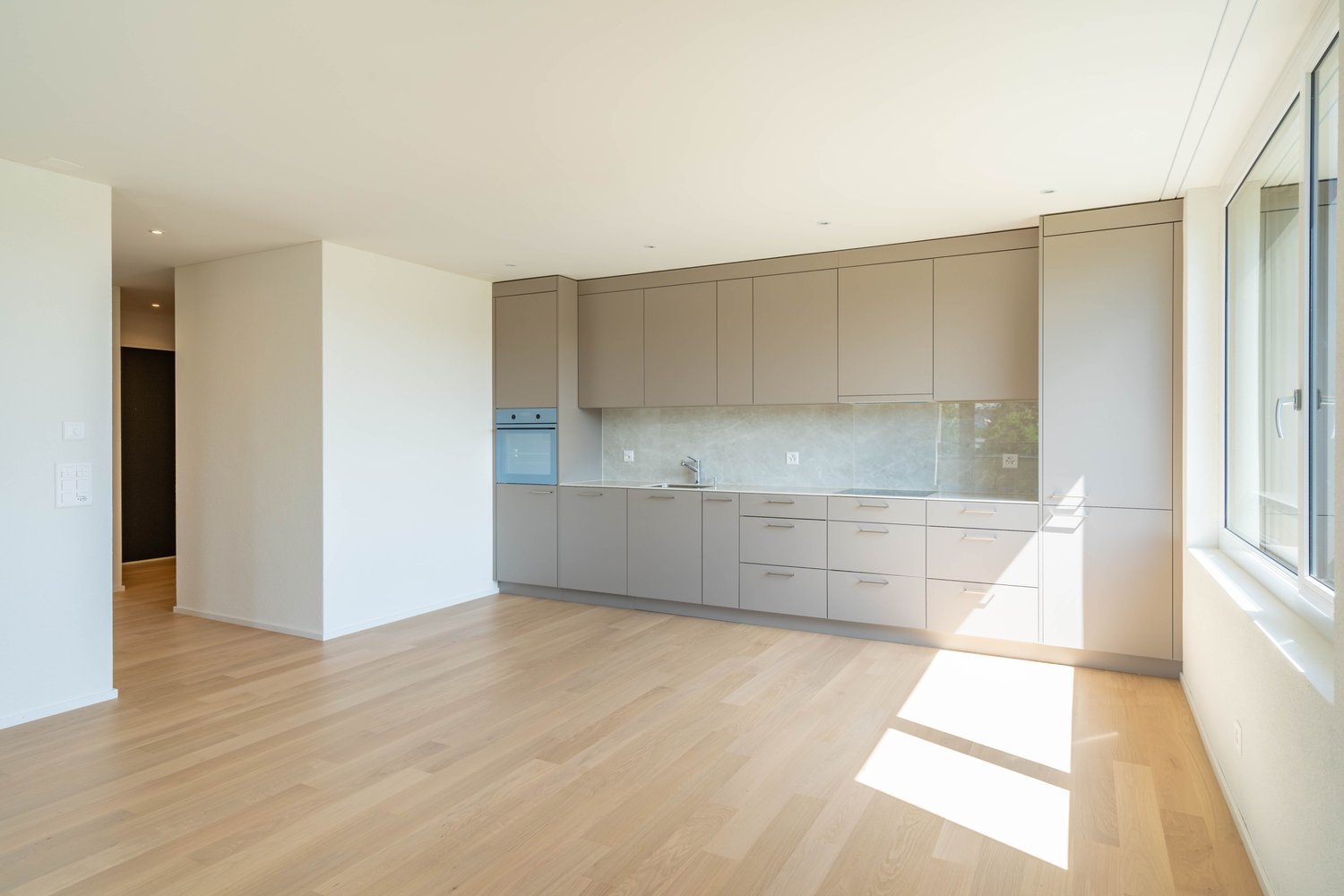 Spacious open-plan kitchen with light wood floors, beige cabinets, built-in appliances including a dishwasher, and large windows providing ample natural light.