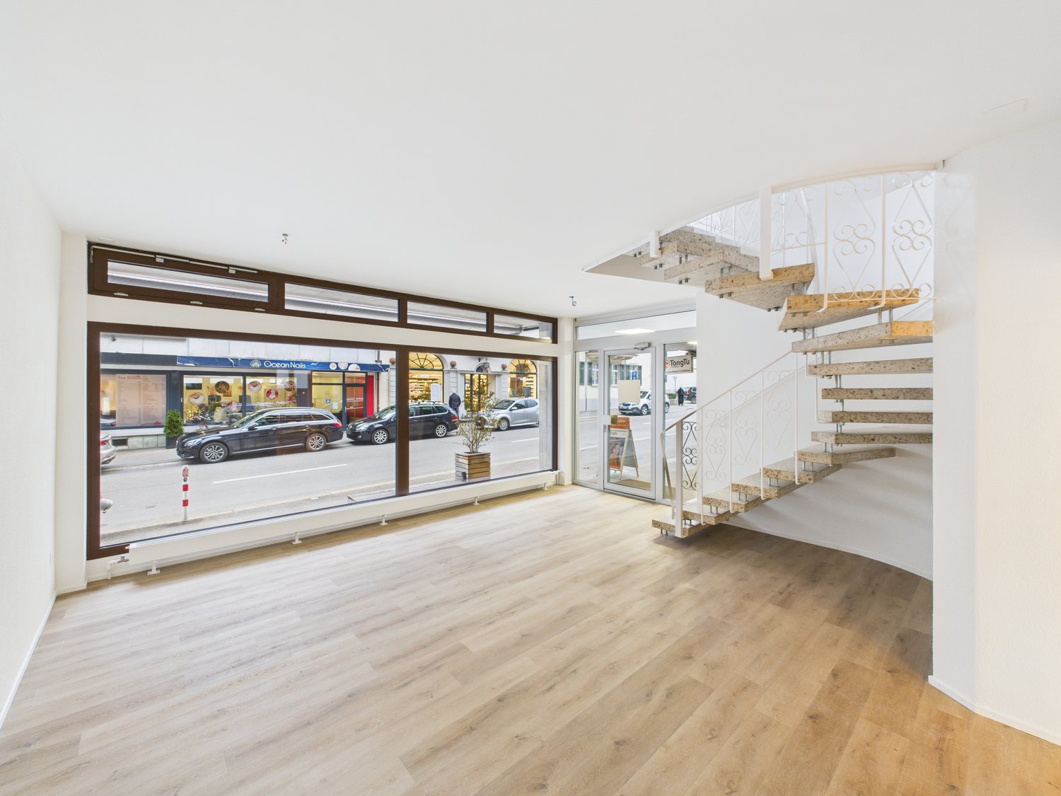 Interior of a commercial space featuring a spacious room with a wooden floor, large windows, a staircase leading to an upper floor, a glass door, a potted plant, and a glass wall.