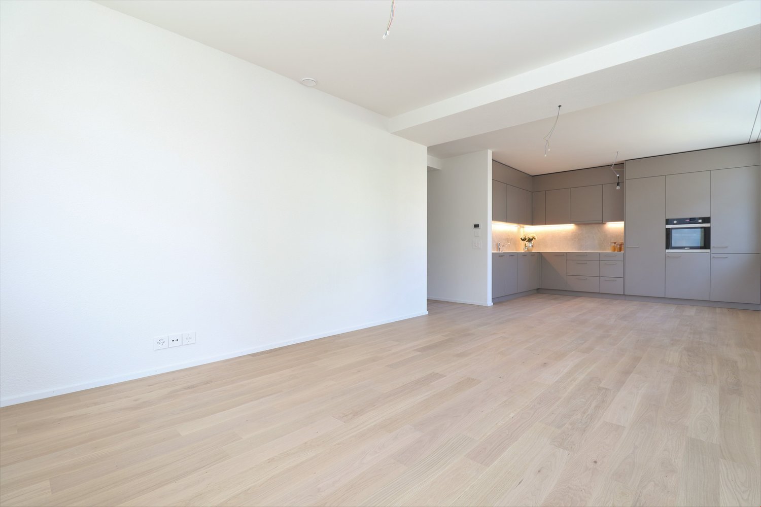 Empty room with wooden floor, white walls, kitchen with cabinets, microwave, and drawers, power outlets on the wall.