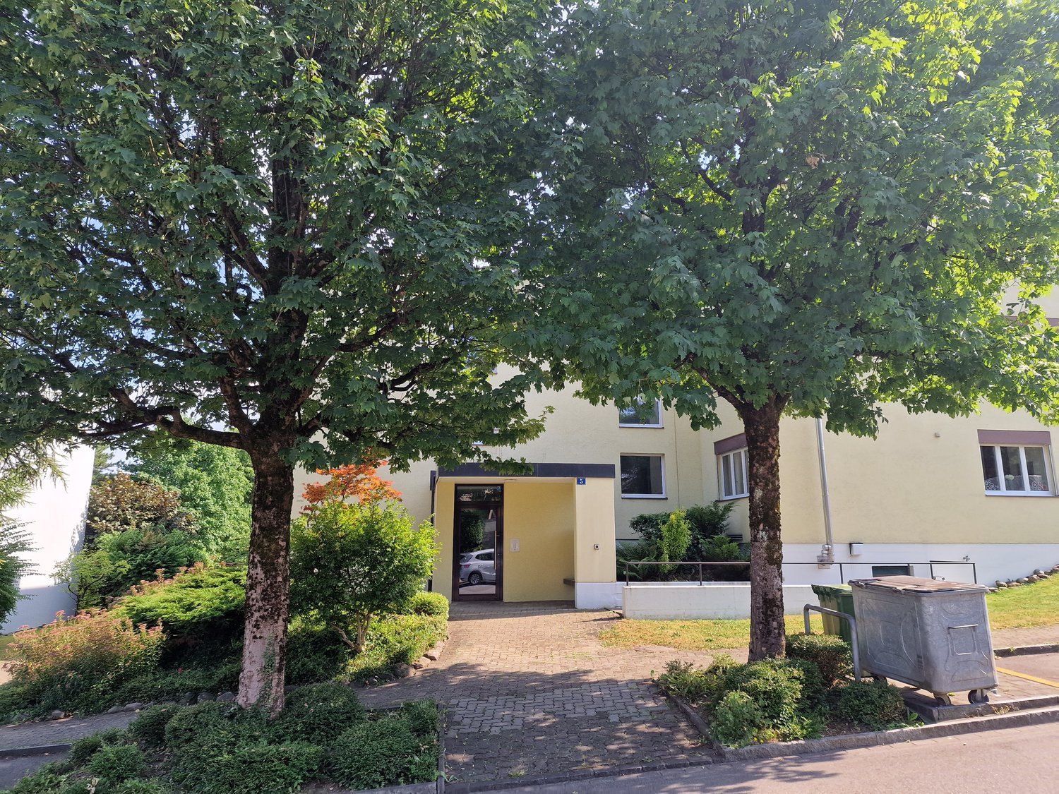 Building with yellow exterior, trash bins near the entrance, paved walkway, shrubs, trees in front, car parked in the doorway
