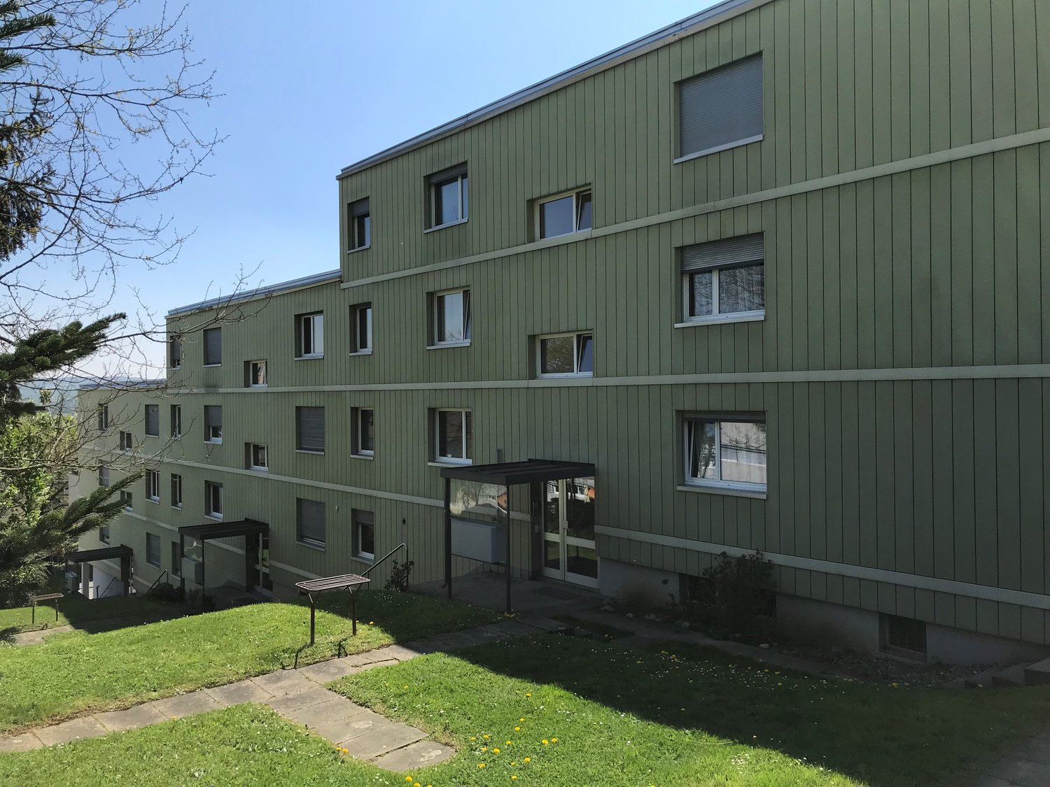 3-story green apartment building with multiple windows, a small grassy area in front, and some trees surrounding the building