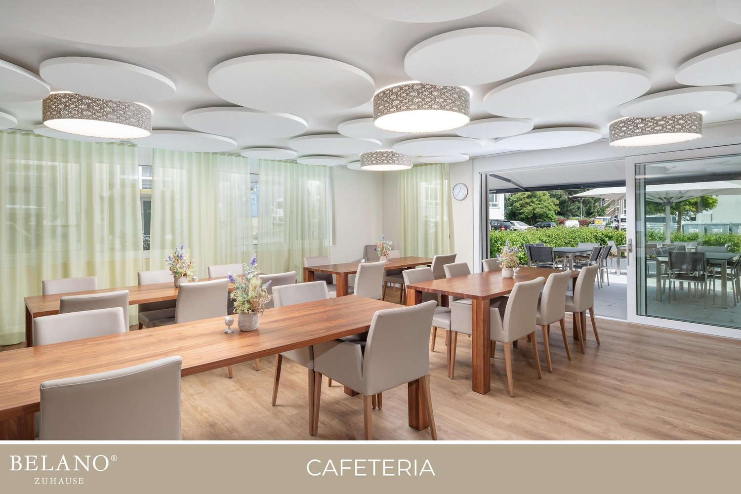Large open dining area with wooden tables and chairs, floor-to-ceiling windows providing a view of the outdoor greenery, and a modern lighting fixture with circular designs on the ceiling.
