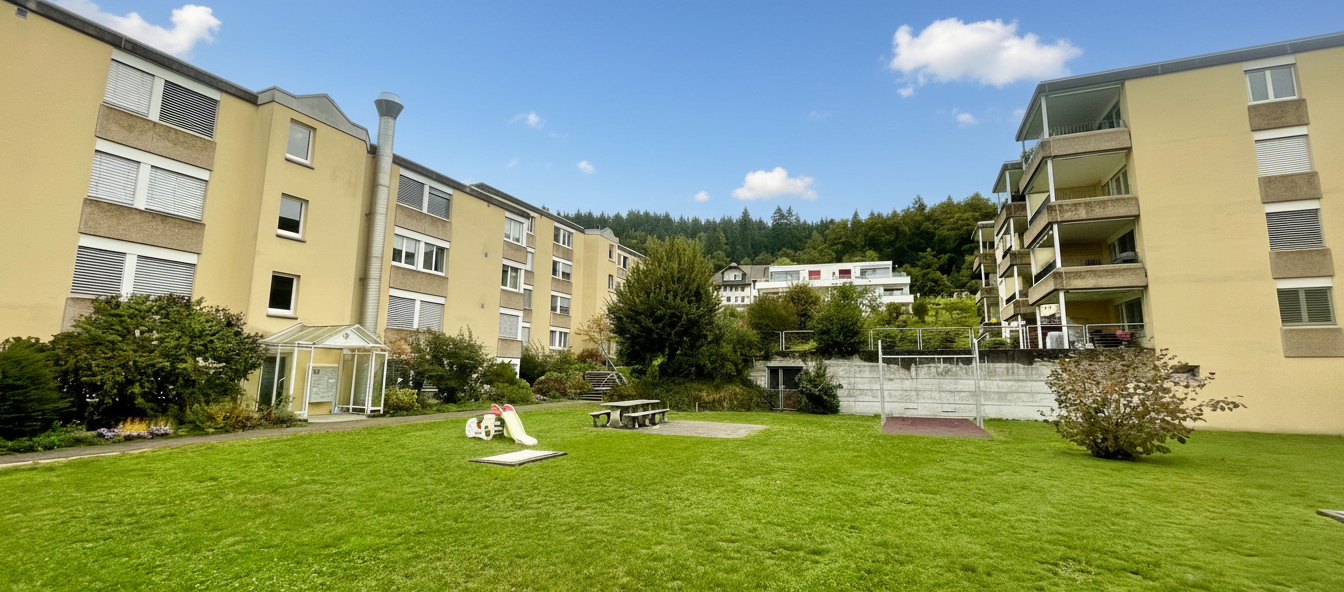 2-story apartment building with several balconies, greenery, slide, benches, fenced backyard with grass