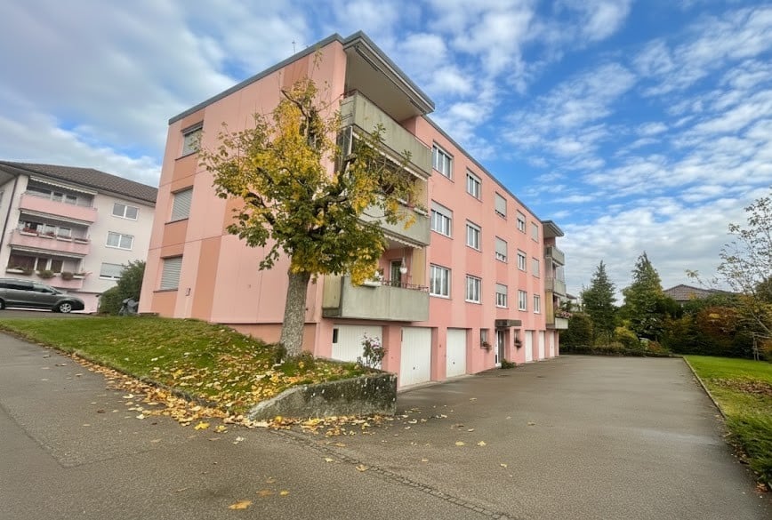 Modern pink building with multiple garages and windows, balcony on some floors, located in a street with parking space