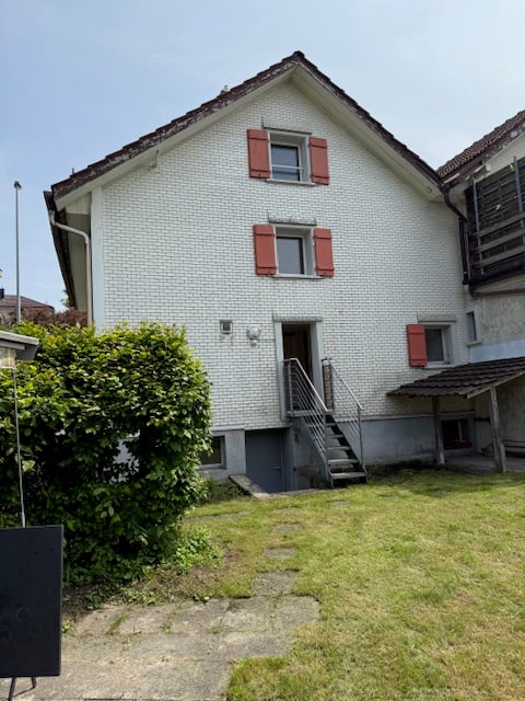 White brick house with red shutters, raised ground floor, stairs, open door, small shed, bushes, green lawn