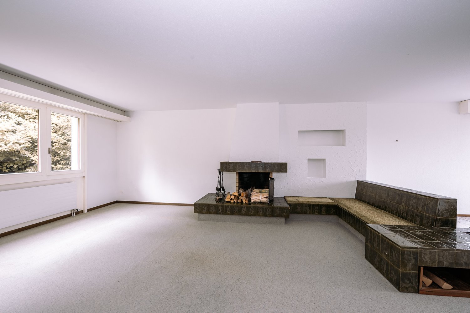 Empty room with white walls, fireplace, large window, beige carpet, built-in benches