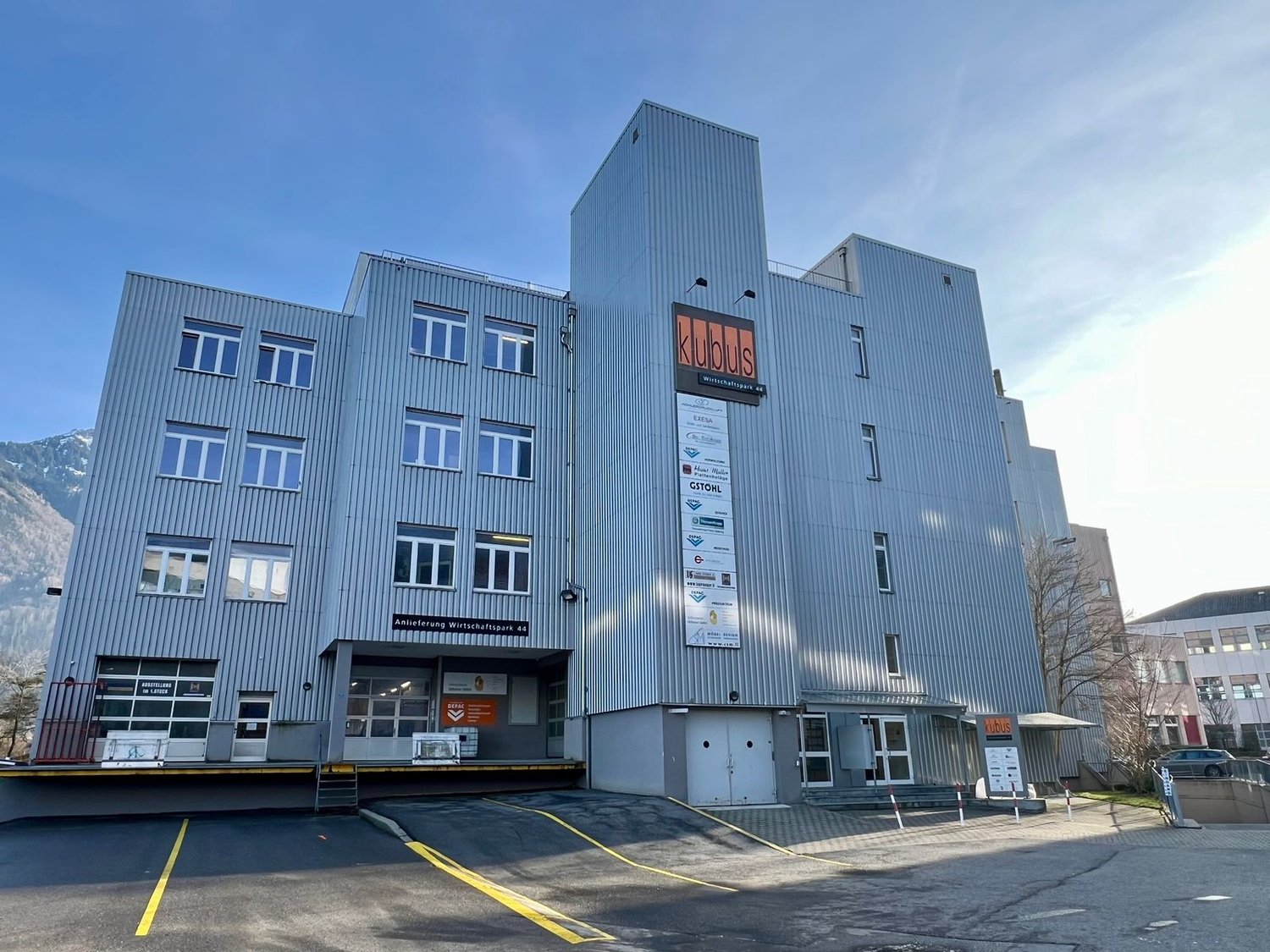 A multi-story commercial building with a gray metal facade and large windows. The building has the name 'Klubus' displayed prominently on the front. There are parking spaces in front of the building.