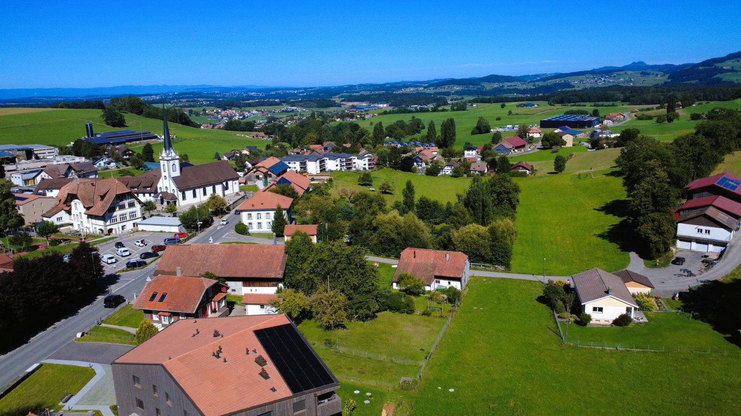 a collection of buildings, green fields, solar panels on roofs, roads, mountains