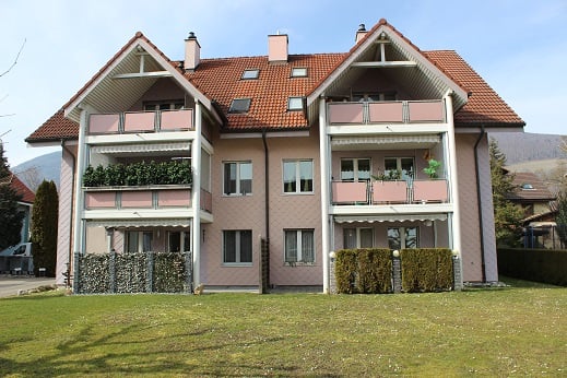 2 story house, red roof, multiple windows, balcony on each floor, large garden, surrounded by hedges, mountains in the background