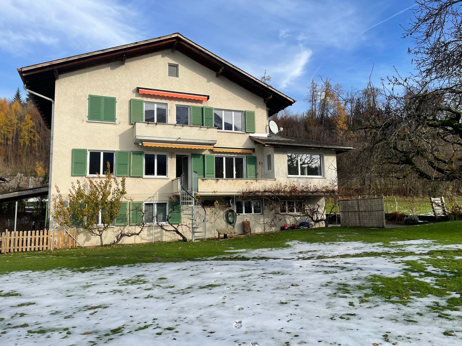 two-story house, green shutters, white windows, satellite dish, white exterior, covered porch, wooden fence, sloped roof, snowy lawn