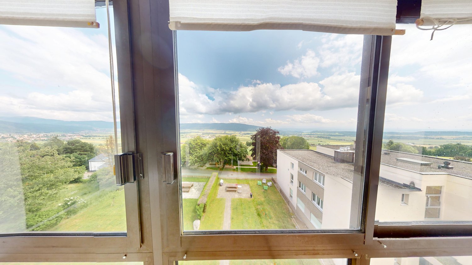 The image shows a view from a window or balcony of an apartment. The view overlooks a green, landscaped area with trees and buildings in the distance. The sky is partly cloudy, creating a pleasant outdoor scene.