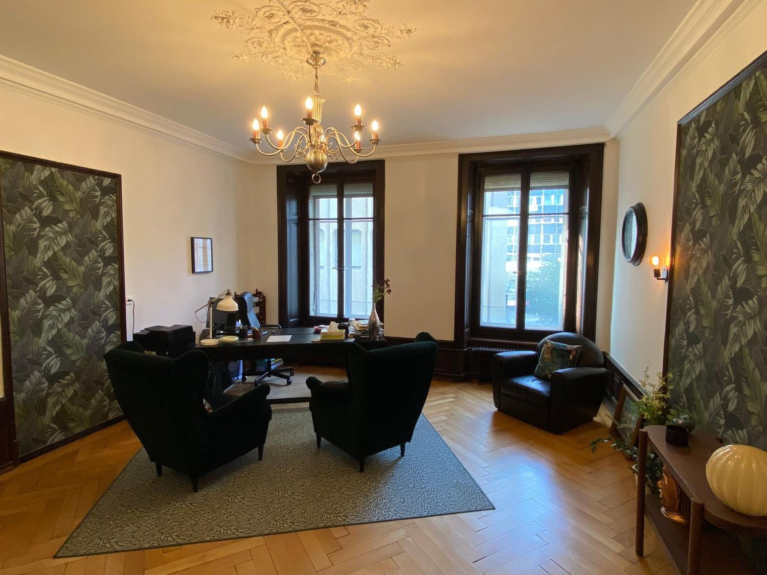 Wooden floor, wooden desk, three black chairs, one armchair, lamps, large windows, chandelier
