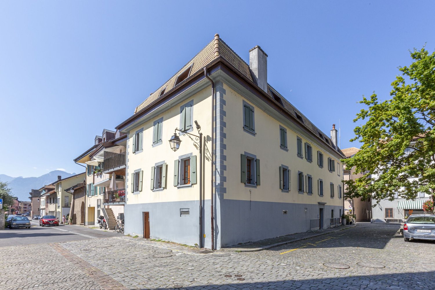 A multi-story building with a slanted roof and stone exterior. The building has several windows and balconies, and is situated on a cobblestone street with other buildings and trees in the background.