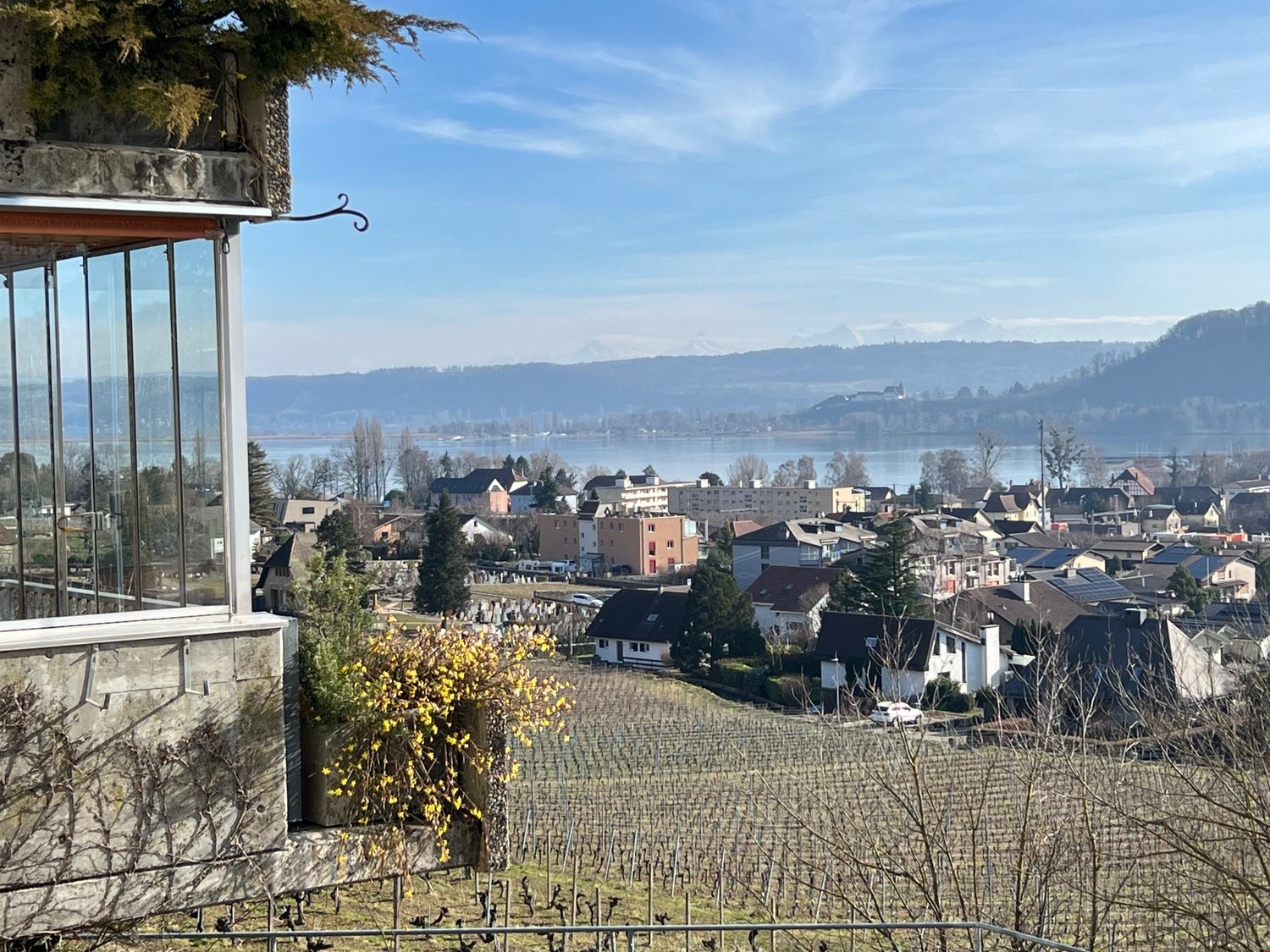 The image shows a scenic view of a town or village nestled in a hilly, mountainous landscape. The town is situated along a body of water, likely a lake or river, with mountains visible in the distance. The foreground features a stone structure with a glas