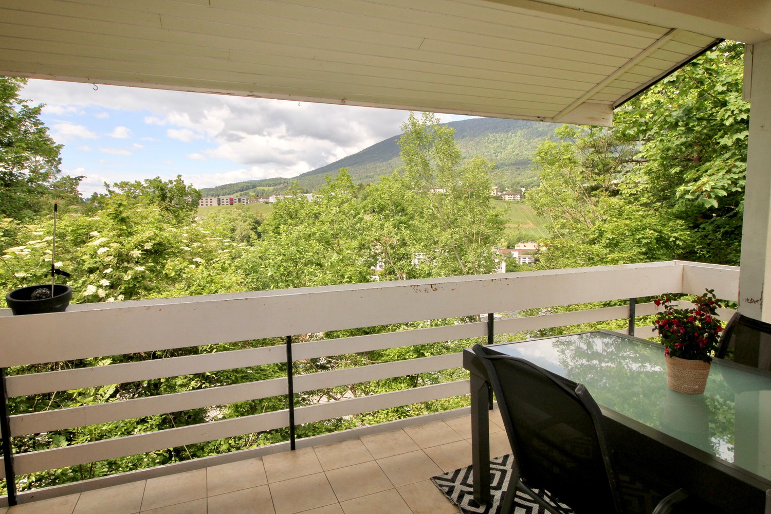 Covered balcony with a glass table and chairs, overlooking a lush, green landscape with mountains in the distance. The balcony is surrounded by trees and vegetation, providing a scenic and peaceful view.