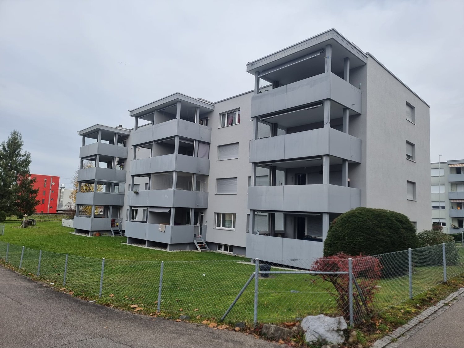 Four story gray apartment building with several balconies, greenery, and fenced yard.