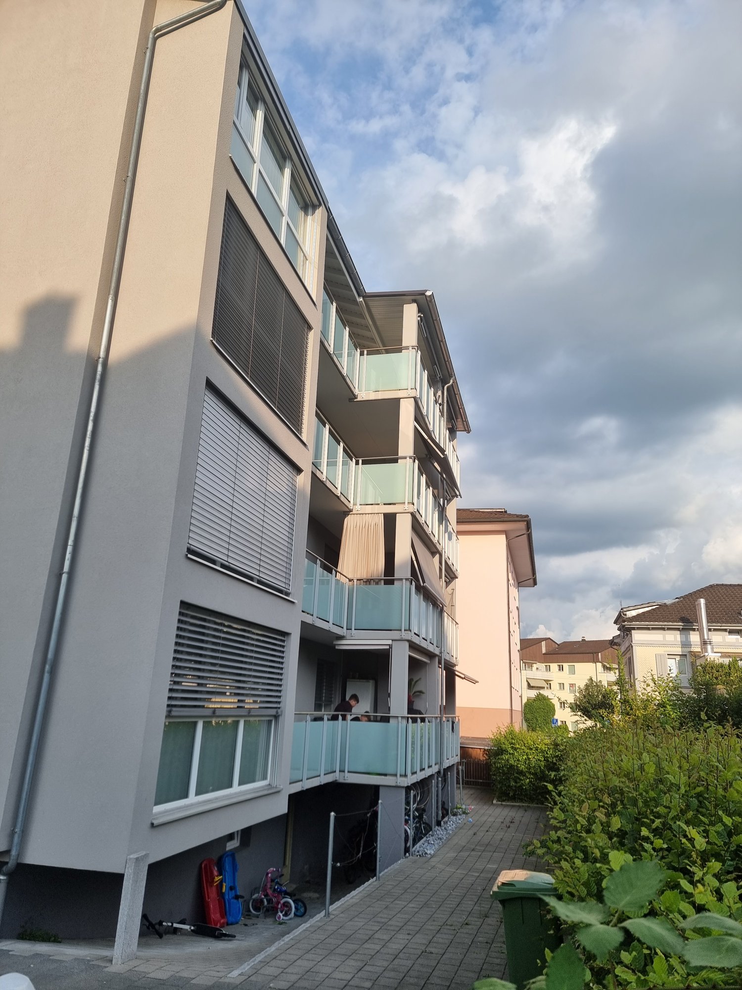 Concrete building with multiple balconies and windows, entrance with a pathway, bikes and strollers