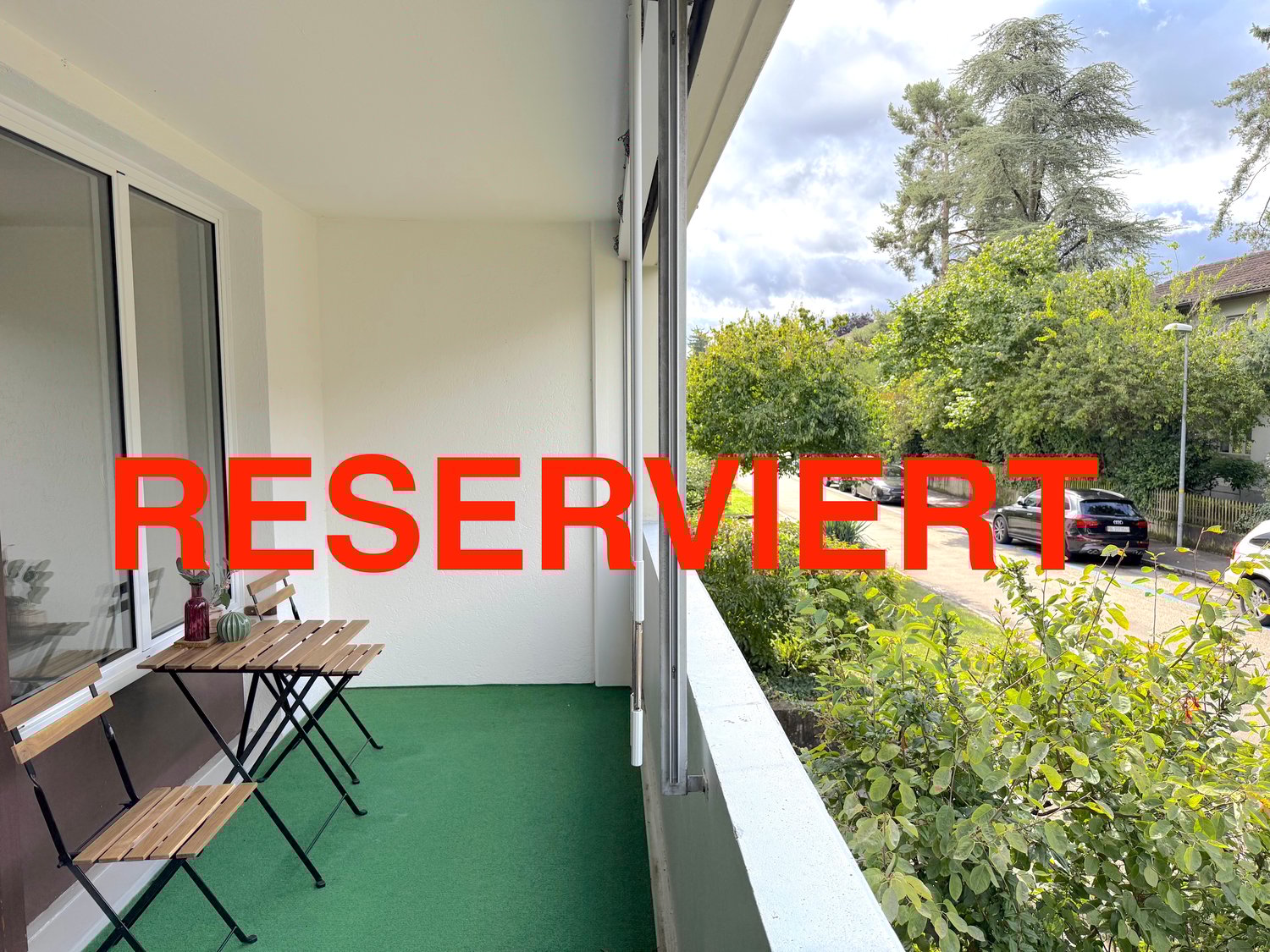 Balcony with a table and chairs, sliding glass door, green carpet, and a view of a tree-lined street.