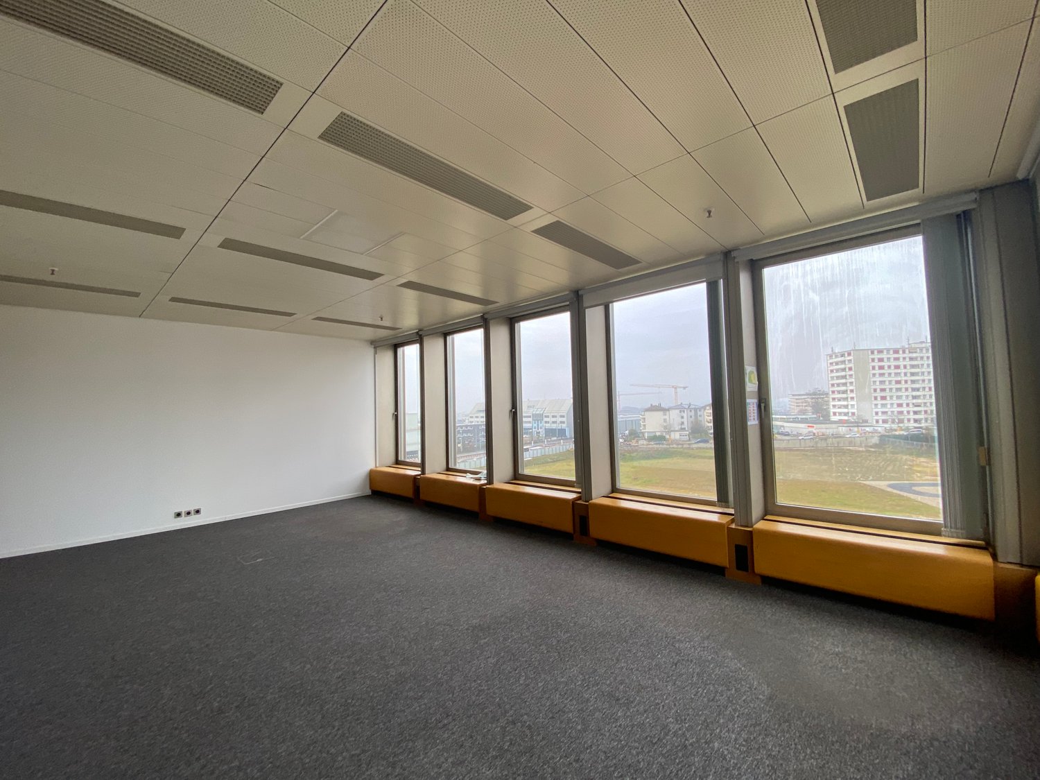 Large open office space with floor-to-ceiling windows providing a view of the surrounding cityscape. The room has a gray carpeted floor and a suspended ceiling with recessed lighting. There are built-in benches along the windows.