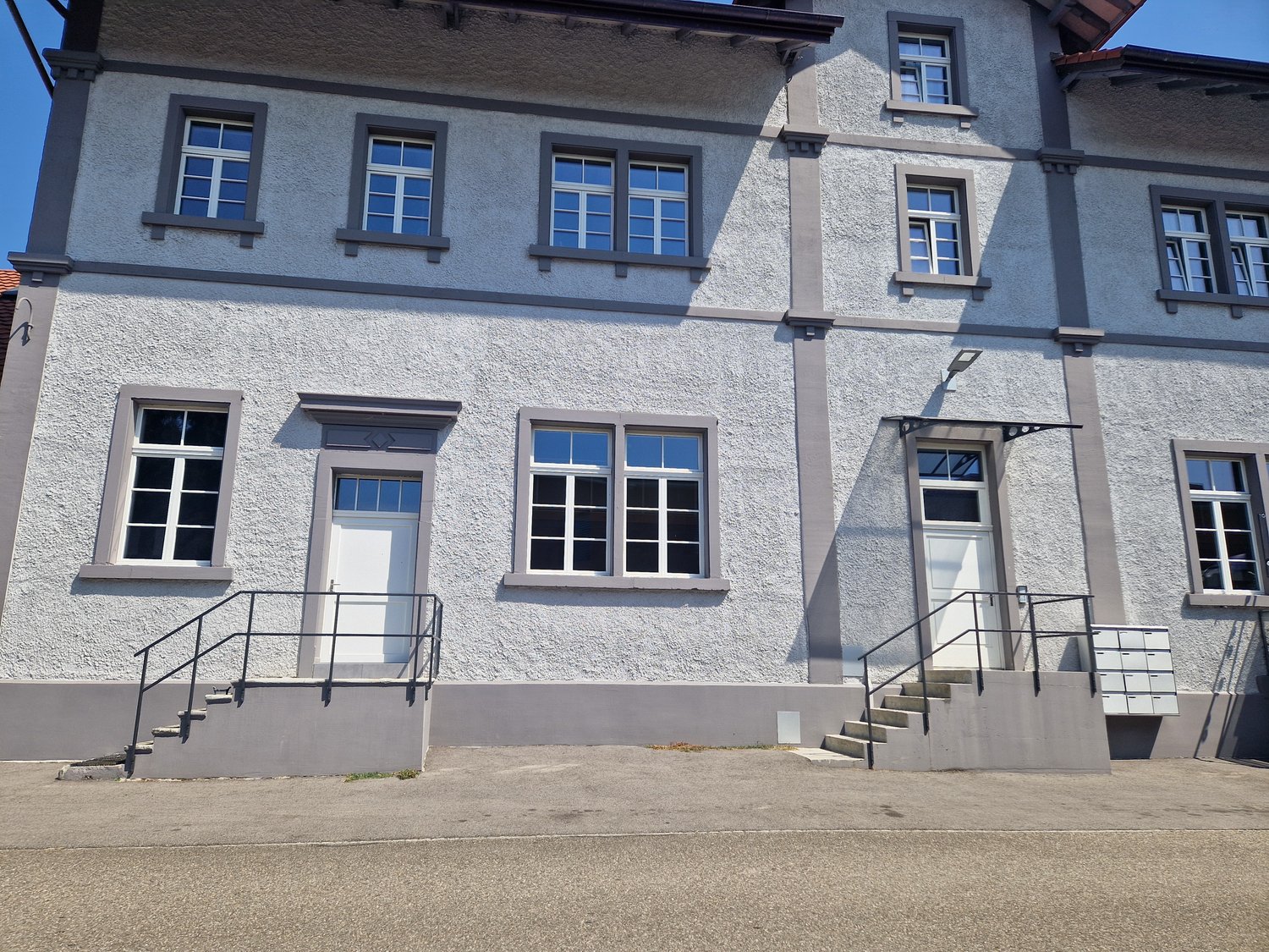 Two-story building, white exterior, glass windows, two entrances with steps, gray handrails, concrete pathway in front