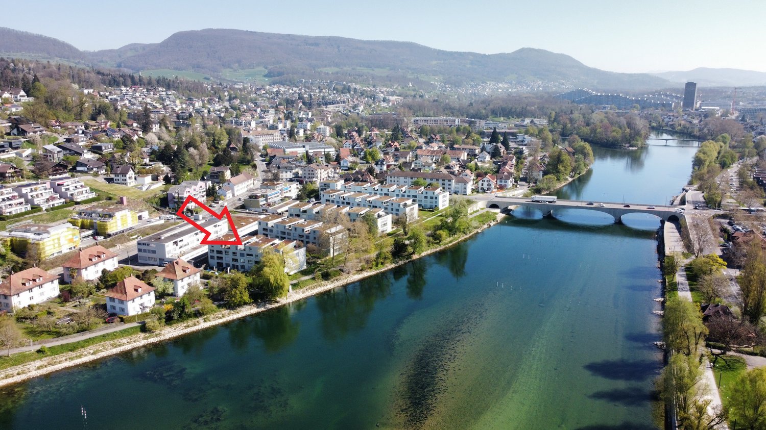 This image shows an aerial view of a town or city situated along a river. The town is surrounded by hills and mountains, with a mix of residential and commercial buildings. There is a bridge crossing the river, and the water appears to be a vibrant blue-g