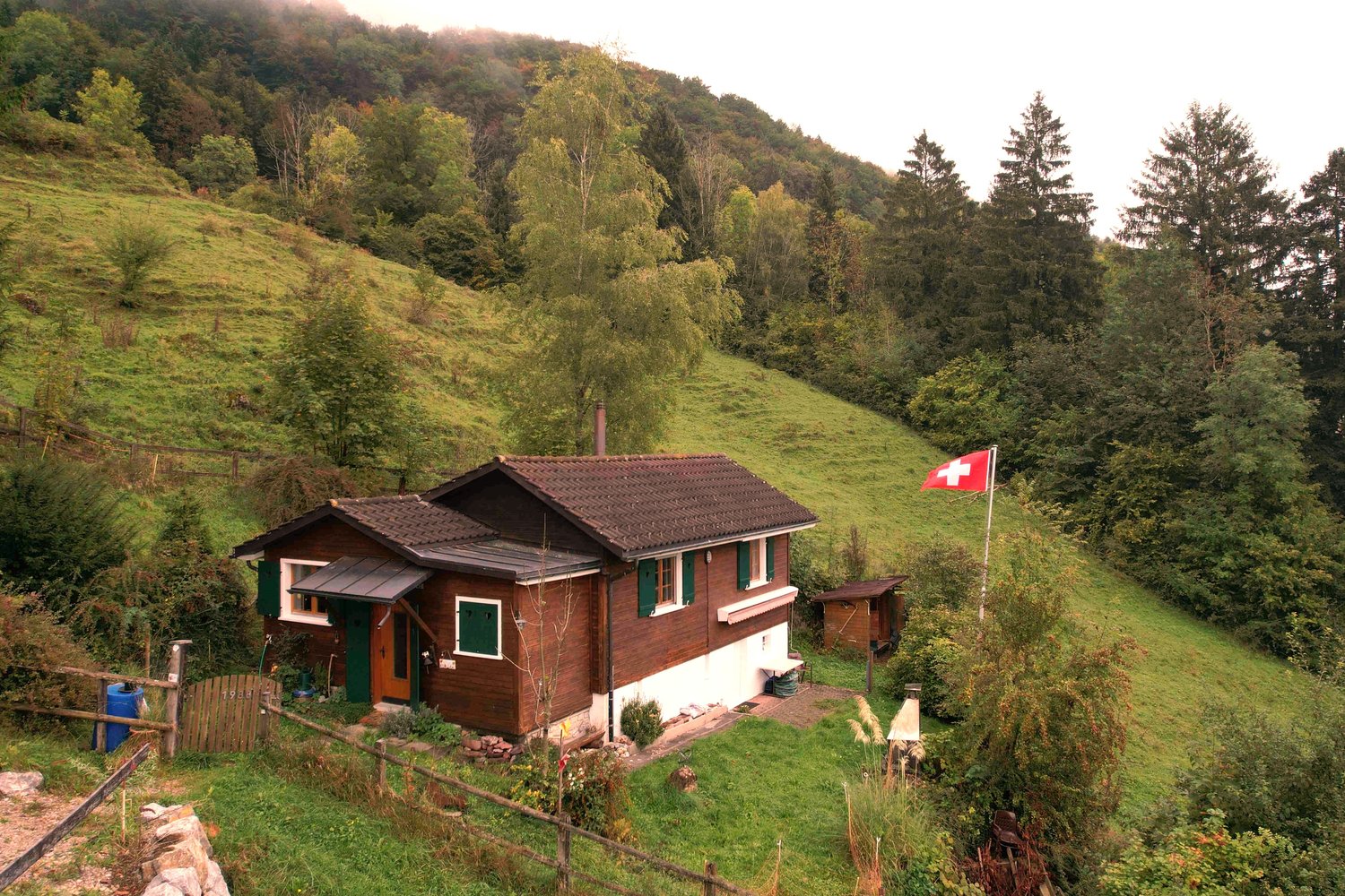Small house on a hill, wooden facade, steep roof, green shutters, fenced yard, Swiss flag, scenic view