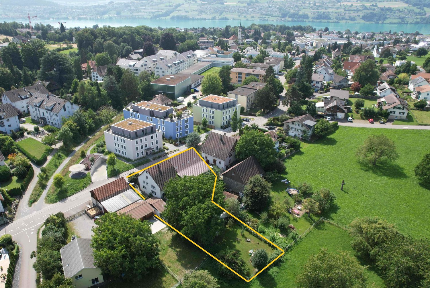 Large residential building, with multiple residential units, surrounded by a large yard with trees and a lake in the background.