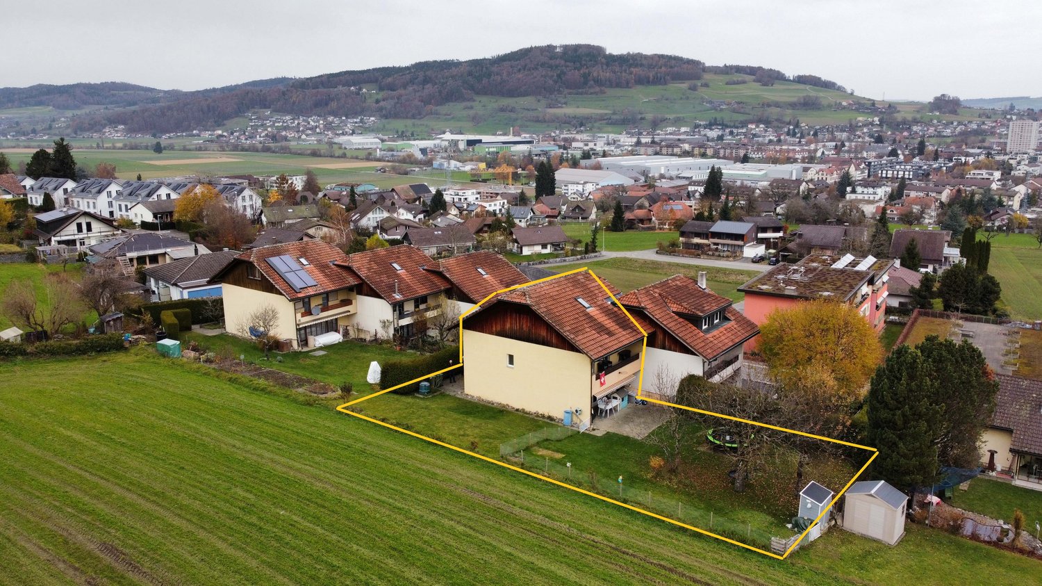 Detached house with solar panels, highlighted plot with a yellow border, green lawns and fields surrounding, road with several parked cars nearby