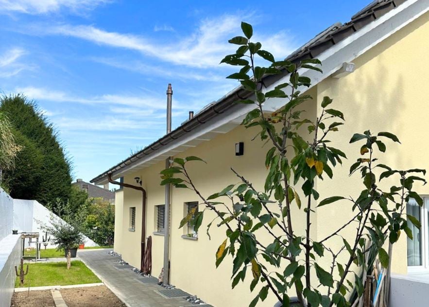 One story house, pale yellow exterior, two visible windows, a patio, a grassy area, a small garden and a concrete walkway.