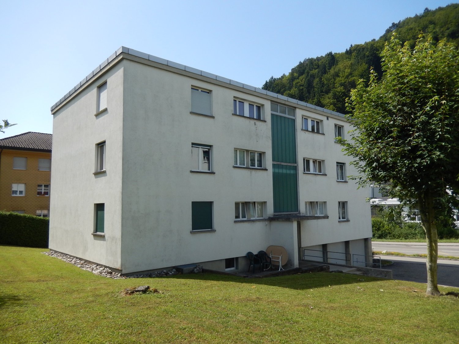 3-story apartment building with a gray exterior, green accents, and large windows. The building is surrounded by trees and has a small parking area in front.