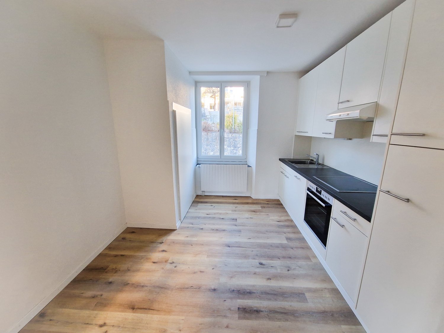 A hallway and a small kitchen in the room, a wooden floor, white walls, white cabinets, black countertops, white ceiling, window, and white radiators.
